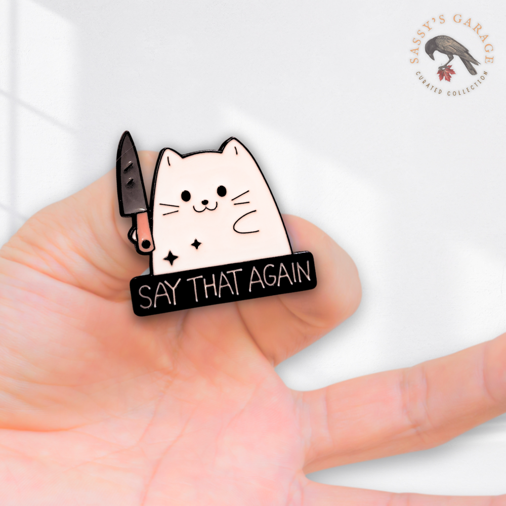 Enamel pin with a cat holding a knife and 'Say That Again' text on a hand.