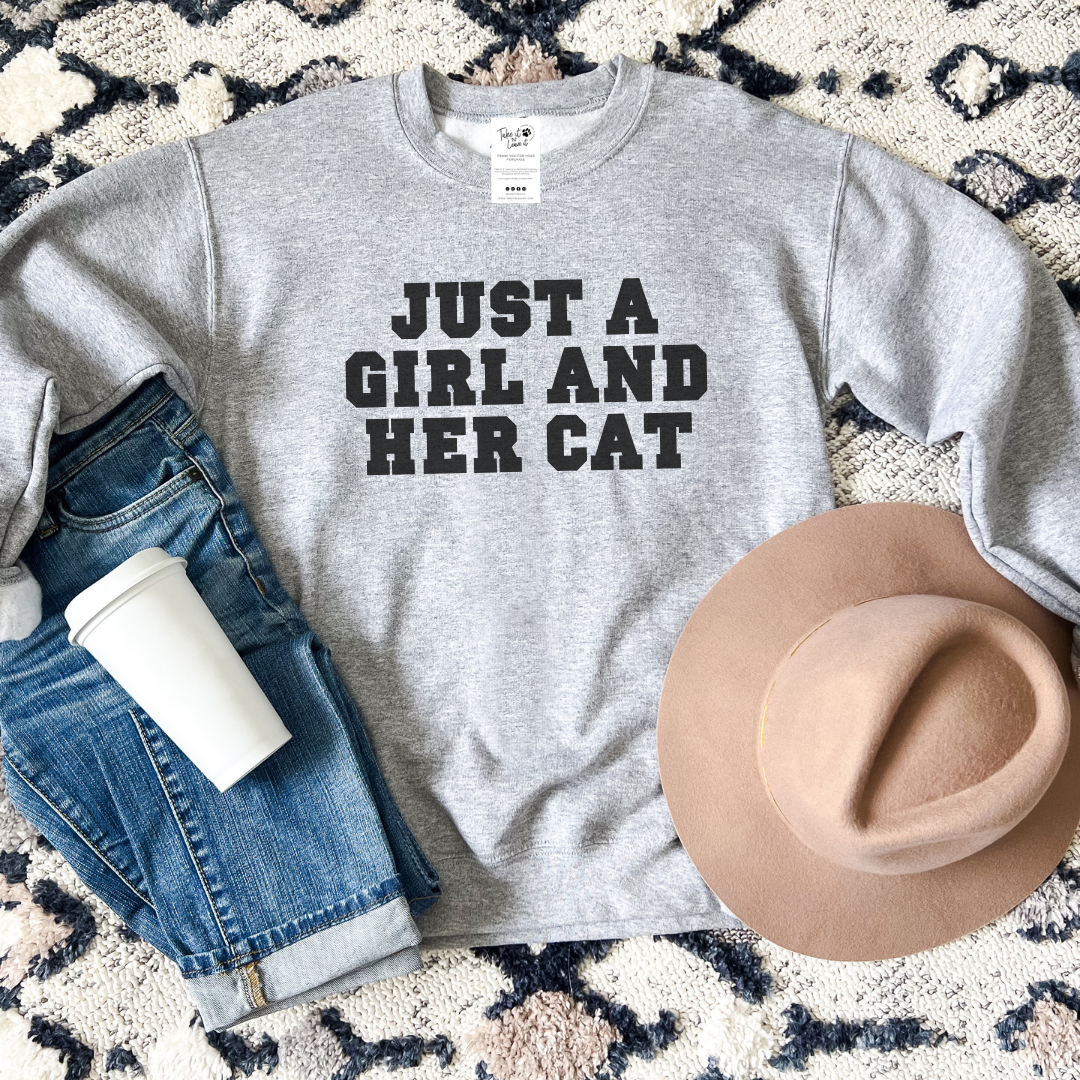 Gray sweatshirt with 'Just a Girl and Her Cat' text, paired with jeans, a hat, and a cup on a patterned surface.
