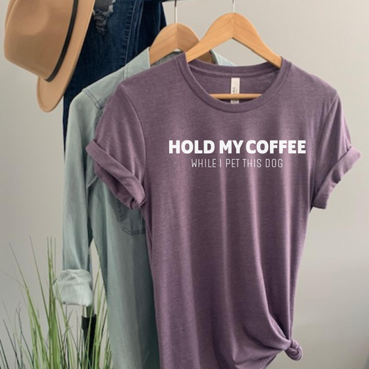 Purple t-shirt with text 'Hold my coffee while I pet this dog' on a hanger against a neutral background.