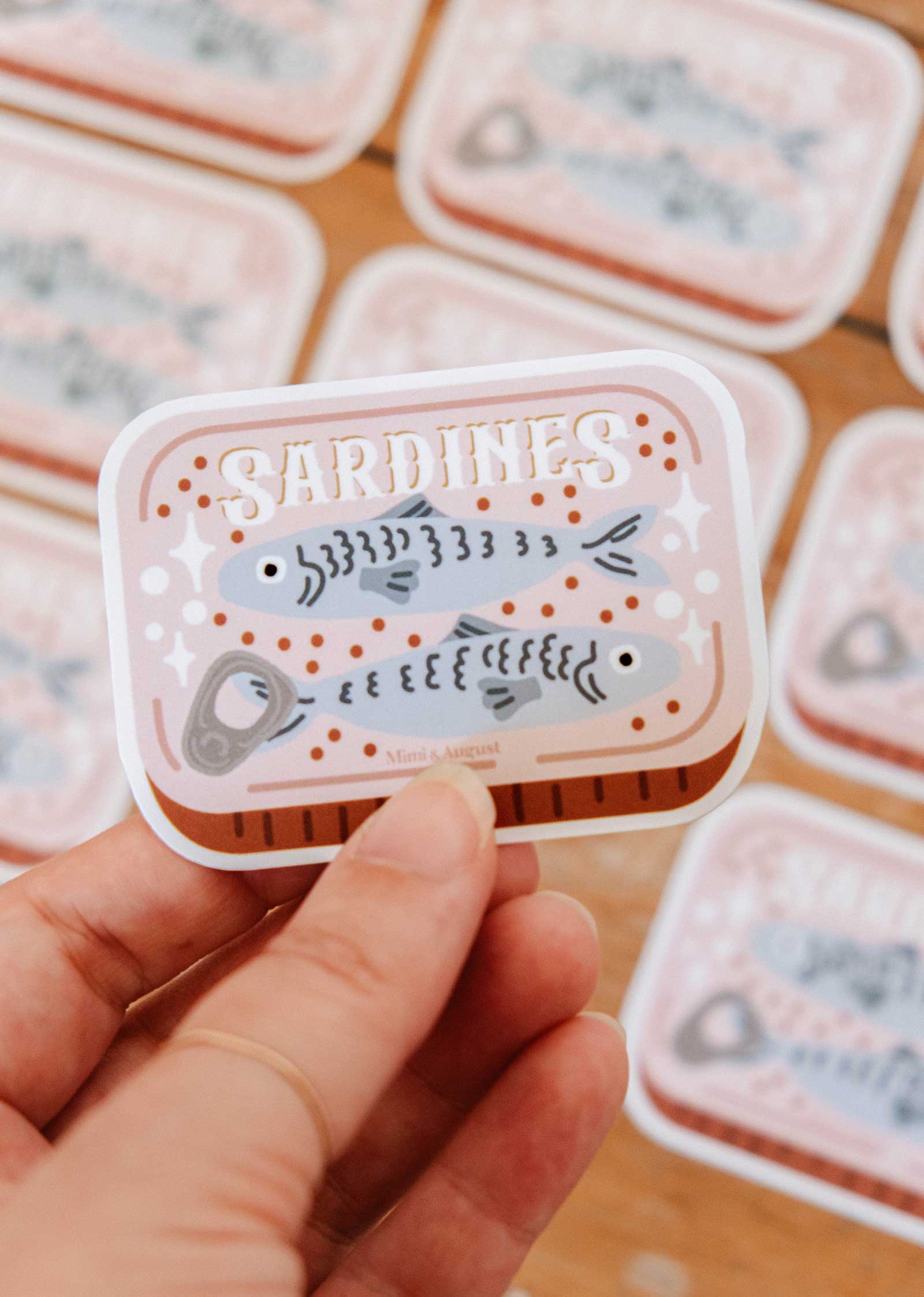 Sticker resembling a sardine can with 'Sardines' text and fish illustrations, held by a hand.